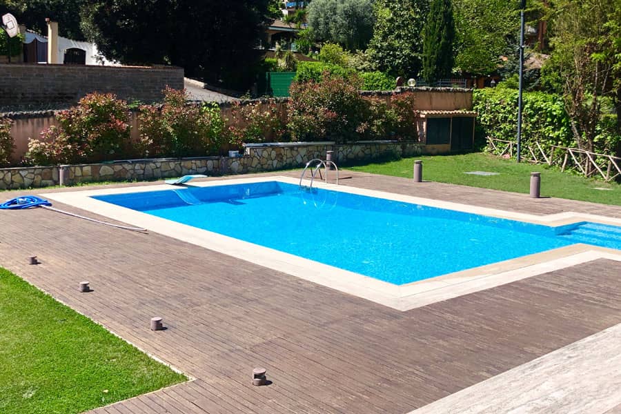 Piscina interrata rettangolare con acqua azzurra, solarium in decking di legno scuro, trampolino e prato circostante in un giardino privato.